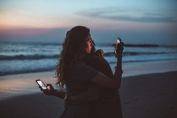 Zonsondergang aan zee. Vrouw en man omhelzen elkaar maar kijken naar hun smartphones.